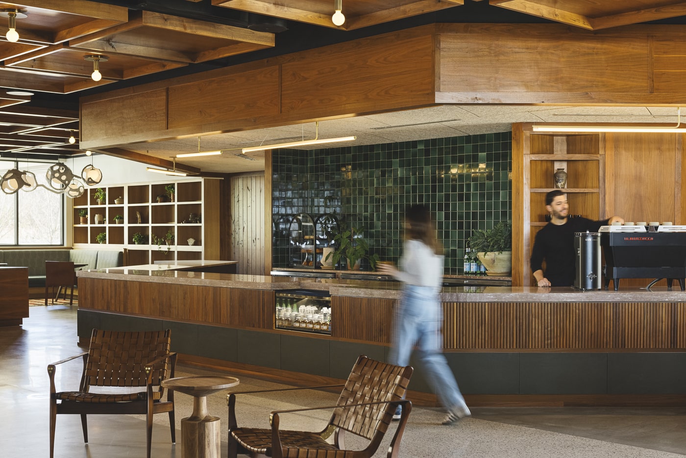 Interior of a modern café or lobby featuring wooden furniture, green tiled walls, and elegant lighting. A barista stands behind a coffee machine, while a blurred figure walks through the space, emphasizing activity and a relaxed atmosphere.
