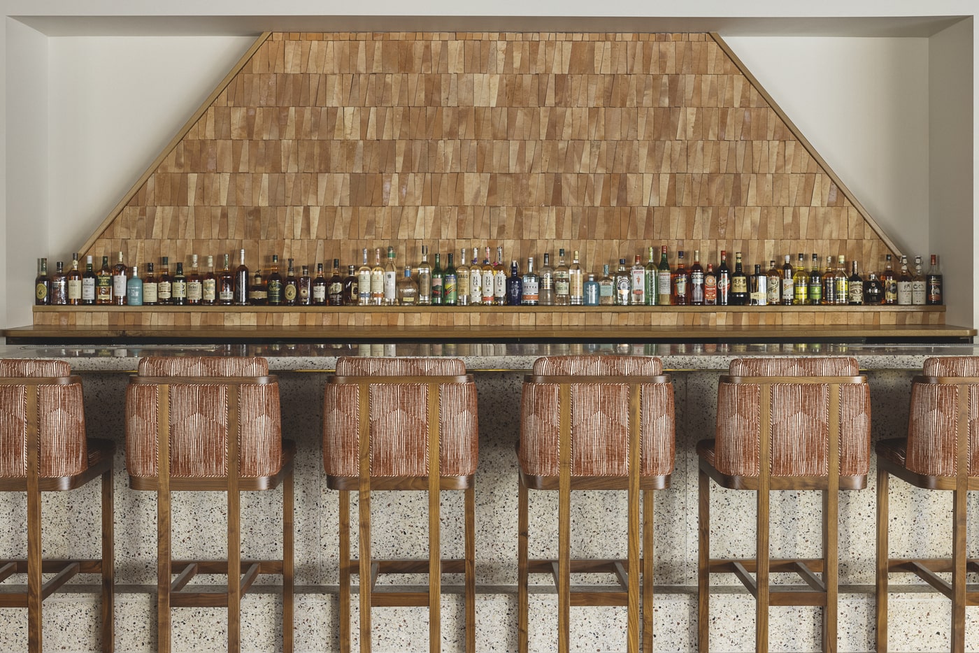 A contemporary bar featuring a geometric wooden backdrop, a sleek arrangement of liquor bottles on a shelf, and stylish upholstered bar stools. The overall design emphasizes symmetry, sophistication, and a warm, inviting atmosphere.
