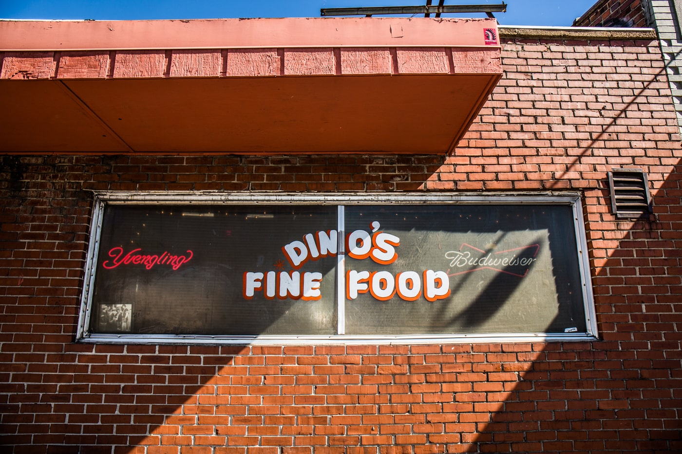 Exterior view of a restaurant or diner with a brick facade featuring a vintage-style window sign that reads "Dino's Fine Food," accompanied by neon signs advertising "Yuengling" and "Budweiser." The image has an urban, nostalgic feel.