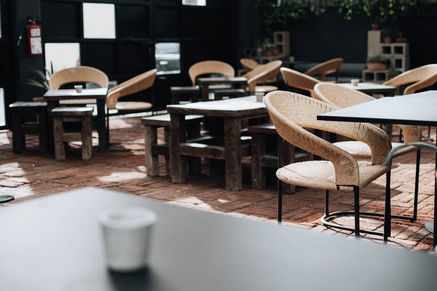 Outdoor café seating area with natural light, featuring wooden tables, woven rattan chairs, and brick flooring. The space has a cozy, rustic vibe with potted plants and minimalistic decor in the background.