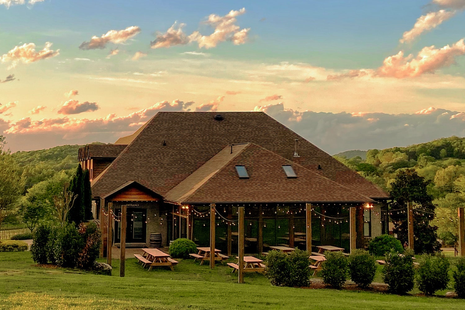A low-slung, brown-shingled building—likely a vineyard tasting room—framed by rolling green hills at sunset.