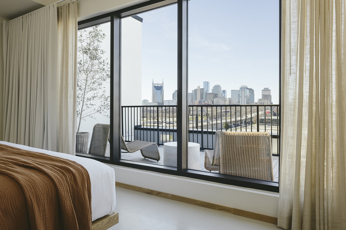 Bright, minimalist hotel room with a white bed draped in a rust-colored blanket at the left edge. 