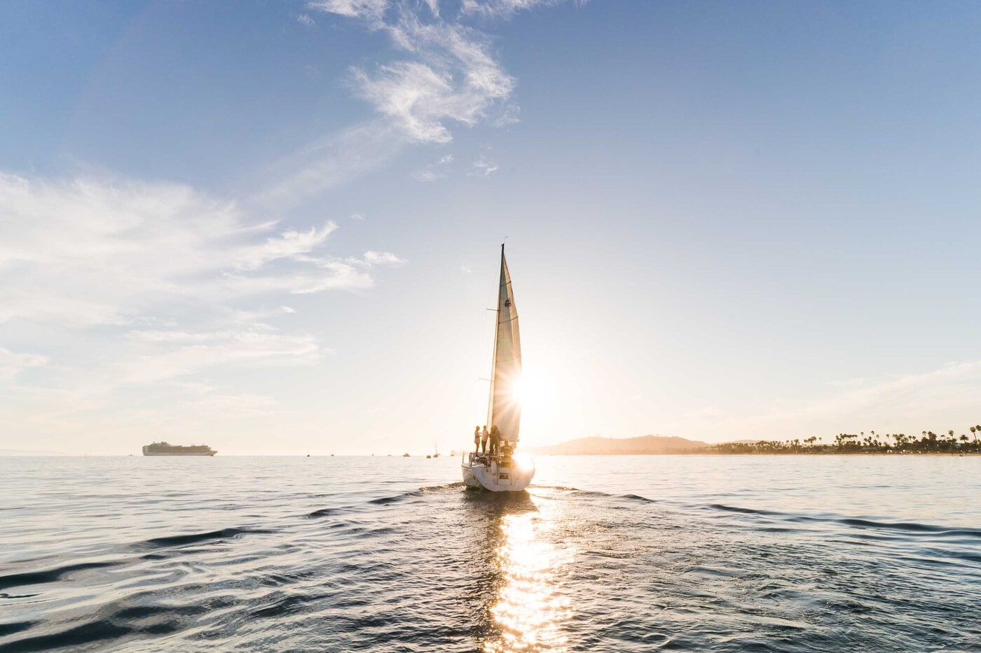 Sailboat with raised mainsail gliding toward a golden sunset over calm, rippling water, passengers gathered at the stern, distant shoreline and palm-lined coast beneath a pale blue sky.