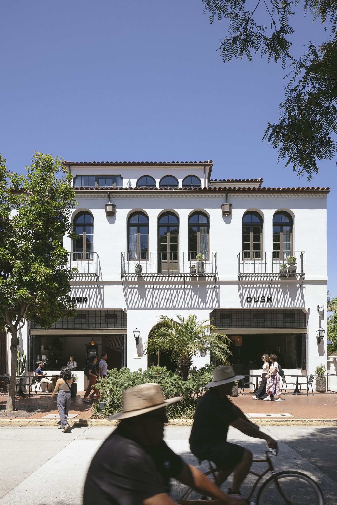 White two-story building with arched windows and iron balconies above open storefronts labeled “Dawn” and “Dusk,” with sidewalk seating, pedestrians and blurred cyclists in the sunlit foreground.