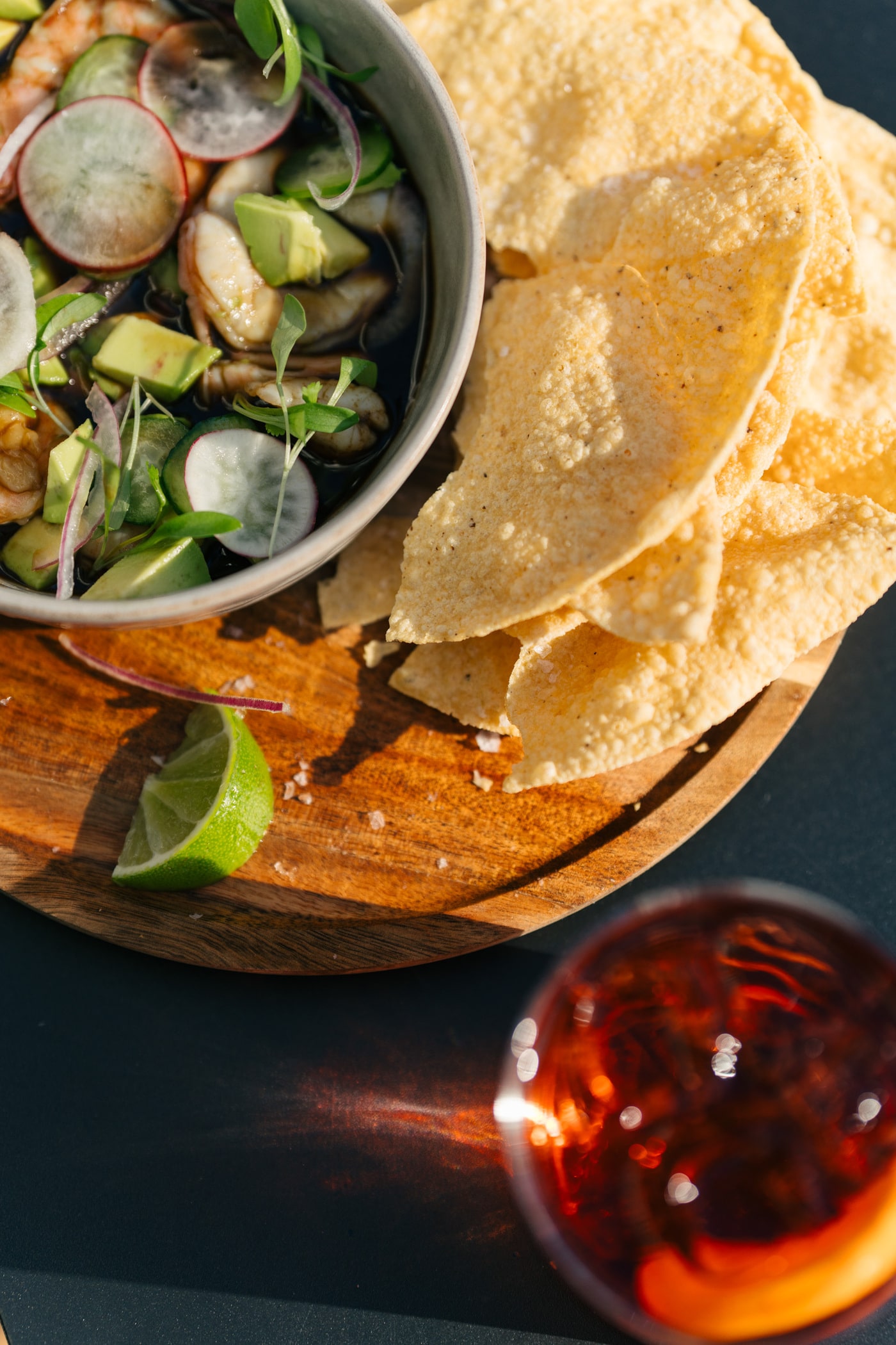 Overhead view of shrimp ceviche topped with radish slices, avocado cubes and microgreens in a bowl beside a stack of tortilla chips, a lime wedge on a wooden board, and a red cocktail glass.