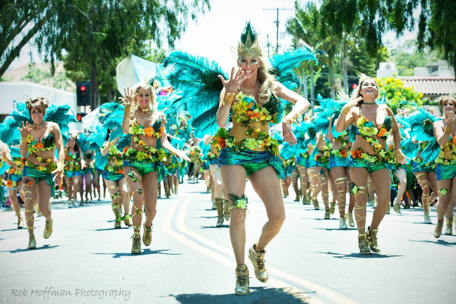 Dancers in green-and-gold costumes with large teal feather wings and floral accents parade down a sunlit street, smiling mid-step in gold sneakers and glittering headpieces.