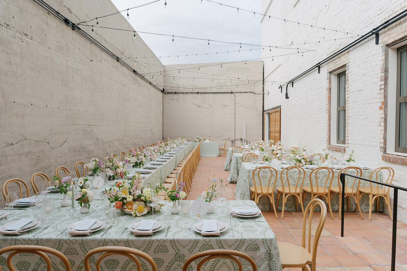 Elegant outdoor wedding setup with long banquet tables, floral arrangements, and string lights in a walled courtyard.