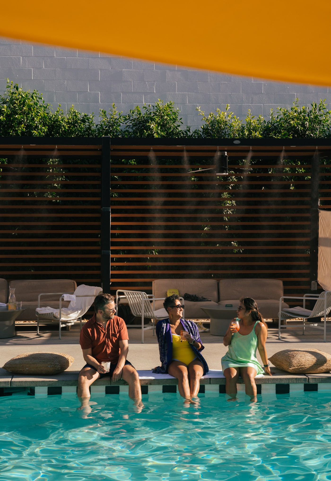 Three friends sit by a sunny poolside, laughing and enjoying drinks, with patio furniture and water mist cooling the area.