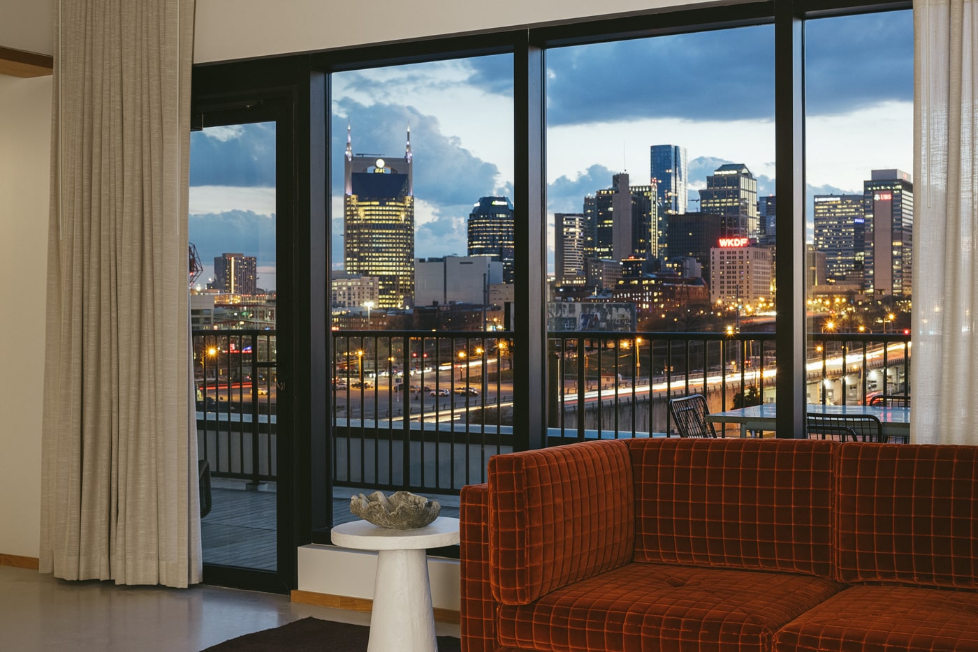 View of a vibrant city skyline at dusk from a modern hotel room, with floor-to-ceiling windows, a red couch, and soft lighting.