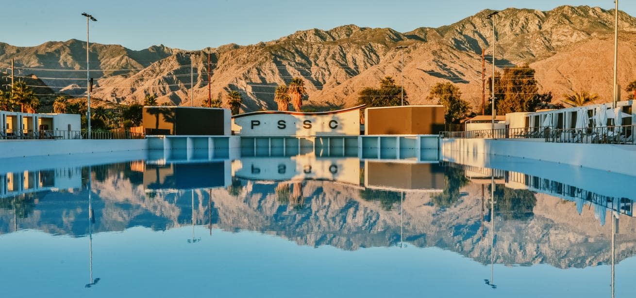 An empty wave pool reflects the surrounding palm trees, buildings, and rugged mountain backdrop under golden sunlight, with "PSSC" letters visible on the central structure.