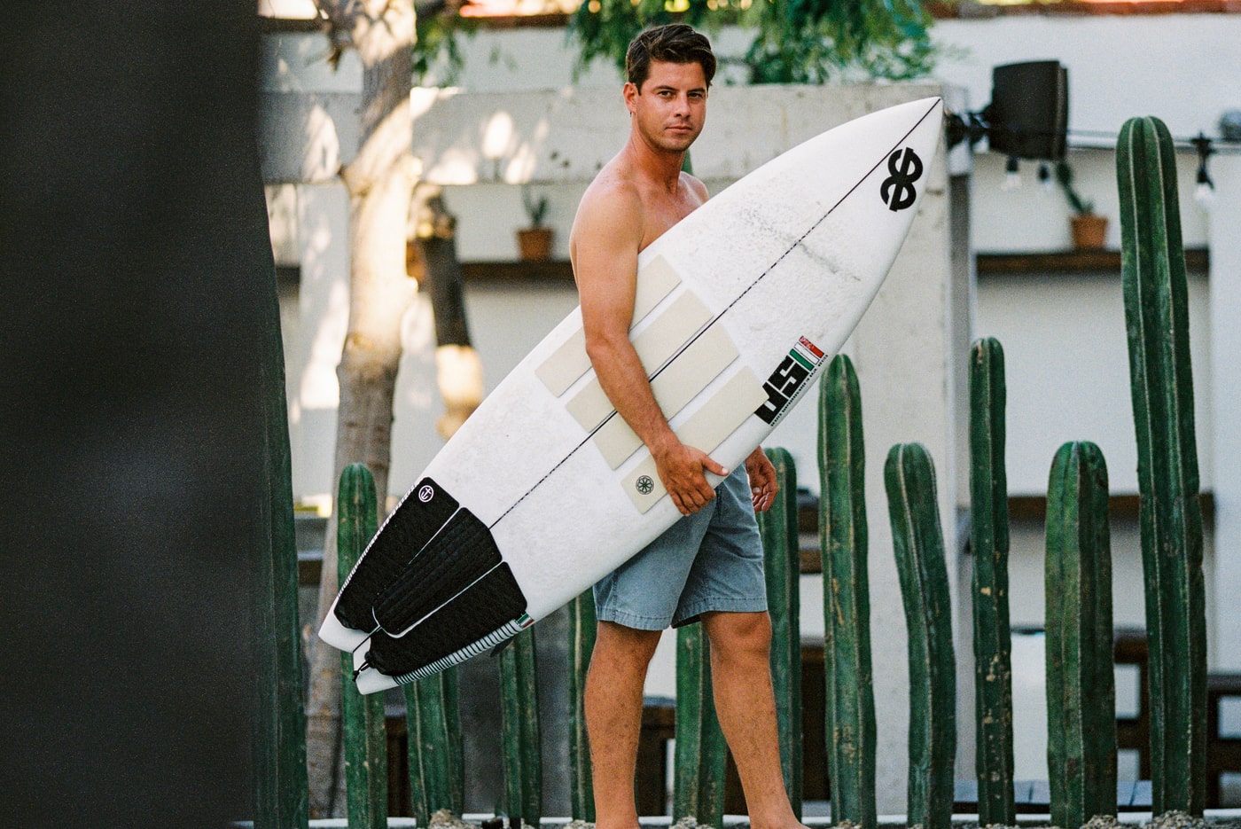 A man stands barefoot holding a surfboard under his arm, wearing gray shorts, with tall green cacti and a white wall in the background.