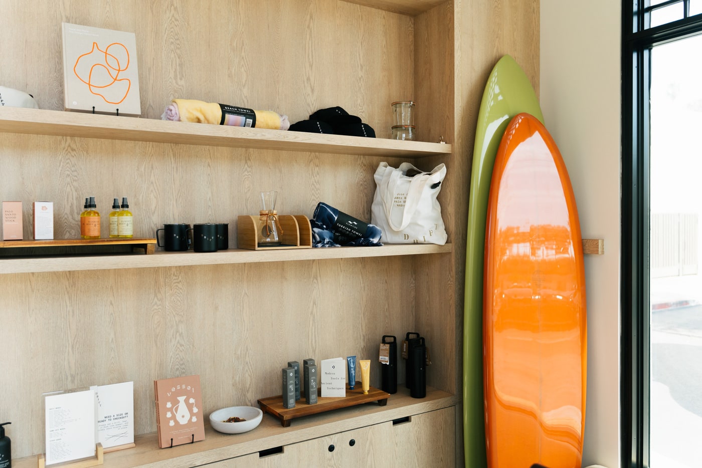 A bright orange and green surfboard stands upright next to wooden shelves stocked with towels, mugs, skincare products, and accessories inside a modern surf shop.