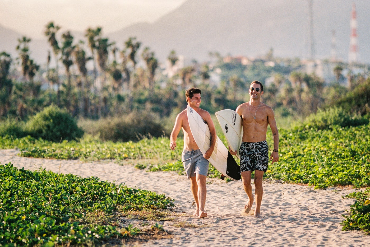 Two smiling surfers walking barefoot on a sandy path toward the beach, carrying surfboards with palm trees and mountains in the background on a sunny day.