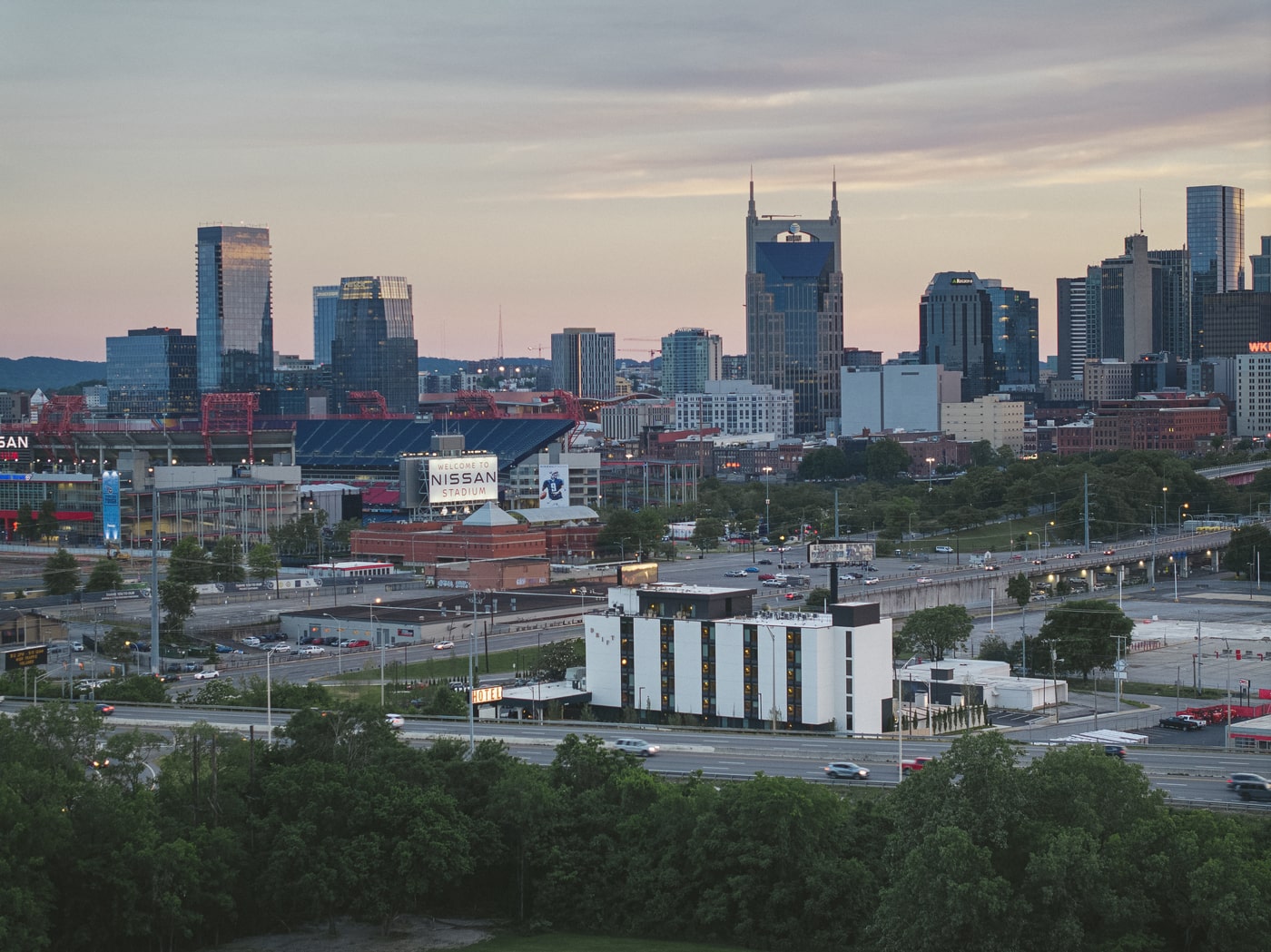 Panoramic view of downtown Nashville at sunset with the iconic AT&T Building, Nissan Stadium, modern skyscrapers, and highways surrounded by trees and city lights.