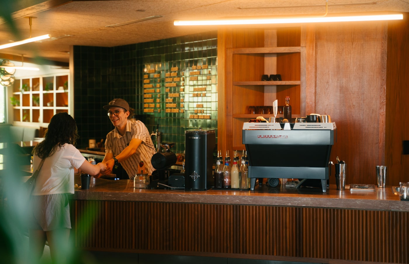 Barista smiling and handing a drink to a customer at a modern café counter with coffee equipment and syrup bottles.