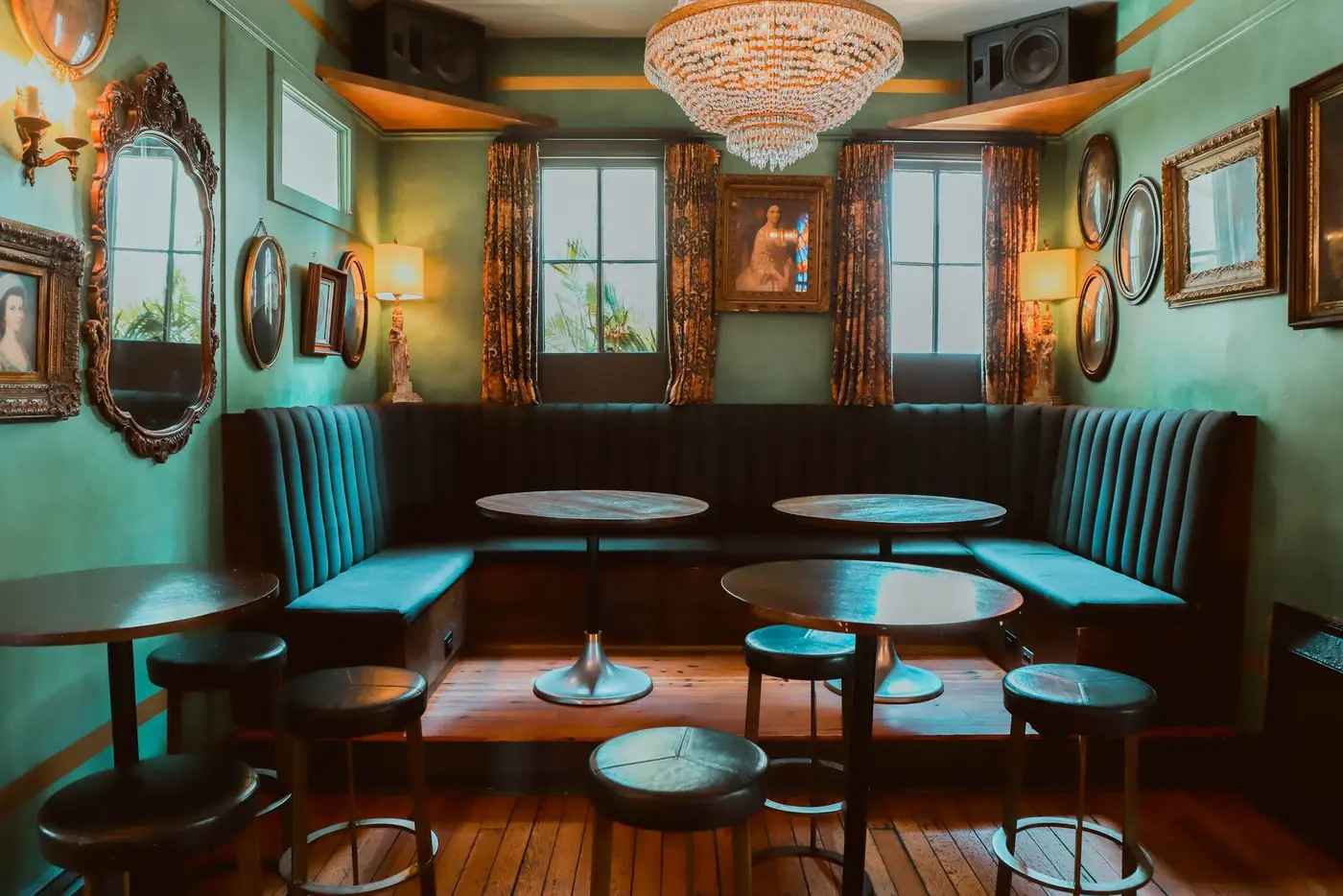 Elegant lounge with teal upholstered seating, round tables, vintage framed art, and a large crystal chandelier under warm lighting.