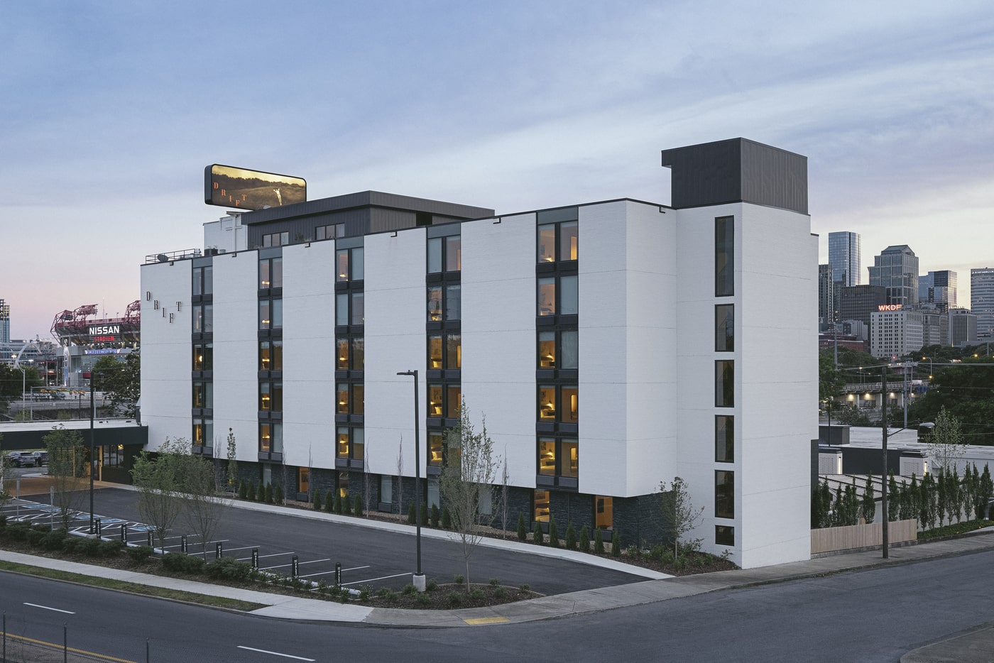 Modern white and black Drift hotel building with large windows, located near Nissan Stadium and downtown Nashville skyline at dusk.