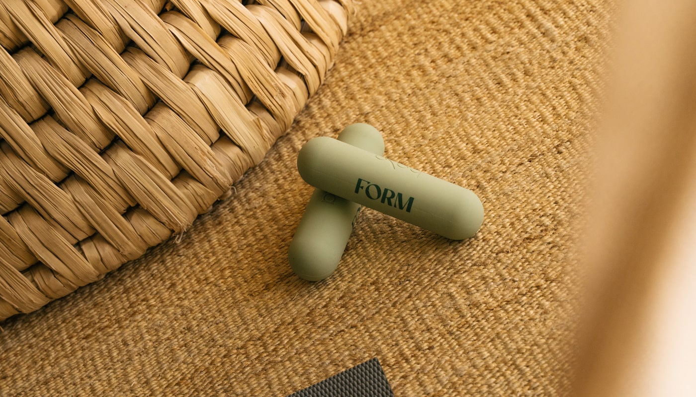 Close-up of green FORM-branded hand weights on a woven mat beside a rattan basket, showcasing minimalist fitness and wellness accessories.