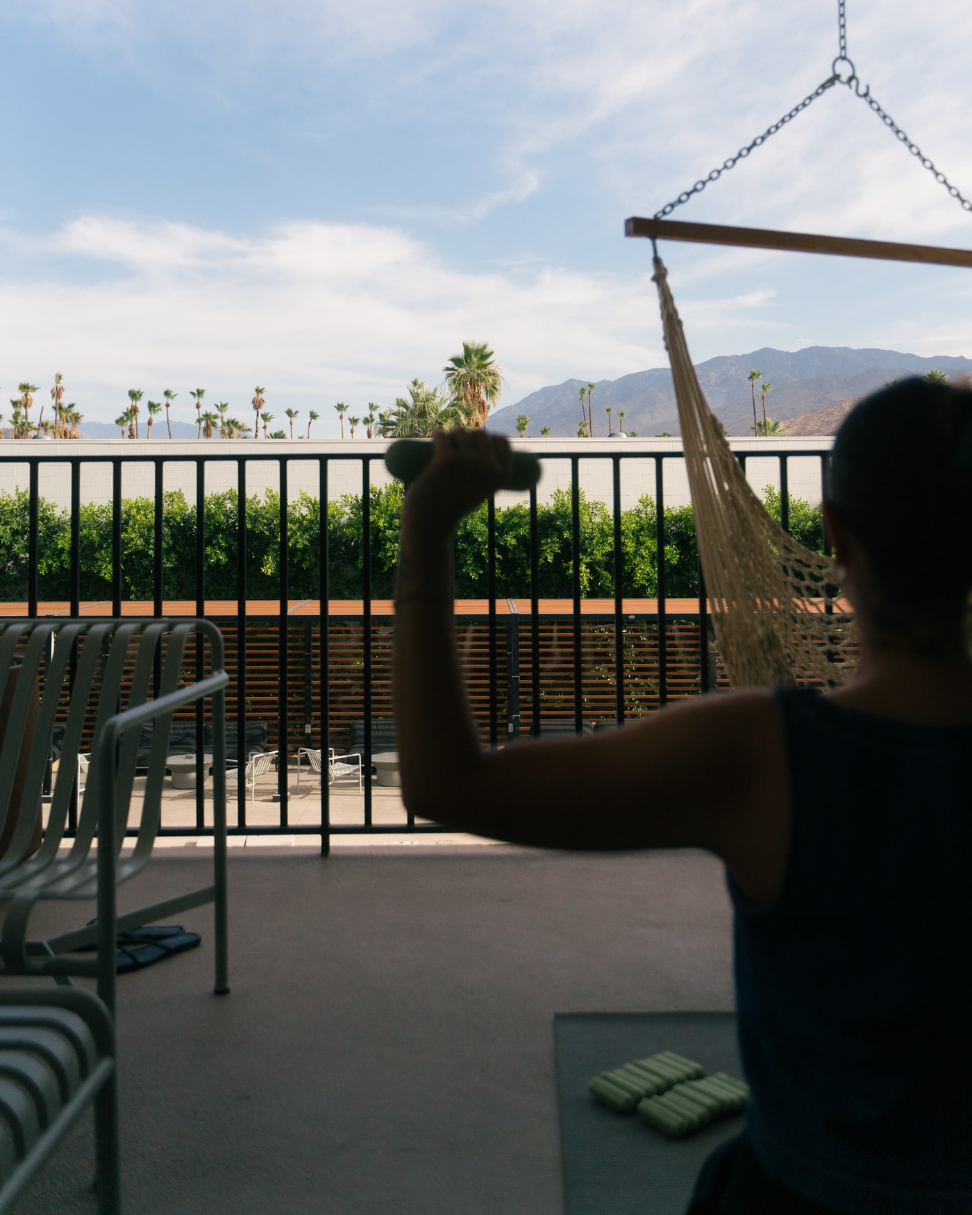 Person exercising on a balcony with a small dumbbell, overlooking palm trees and distant mountains under a partly cloudy sky.