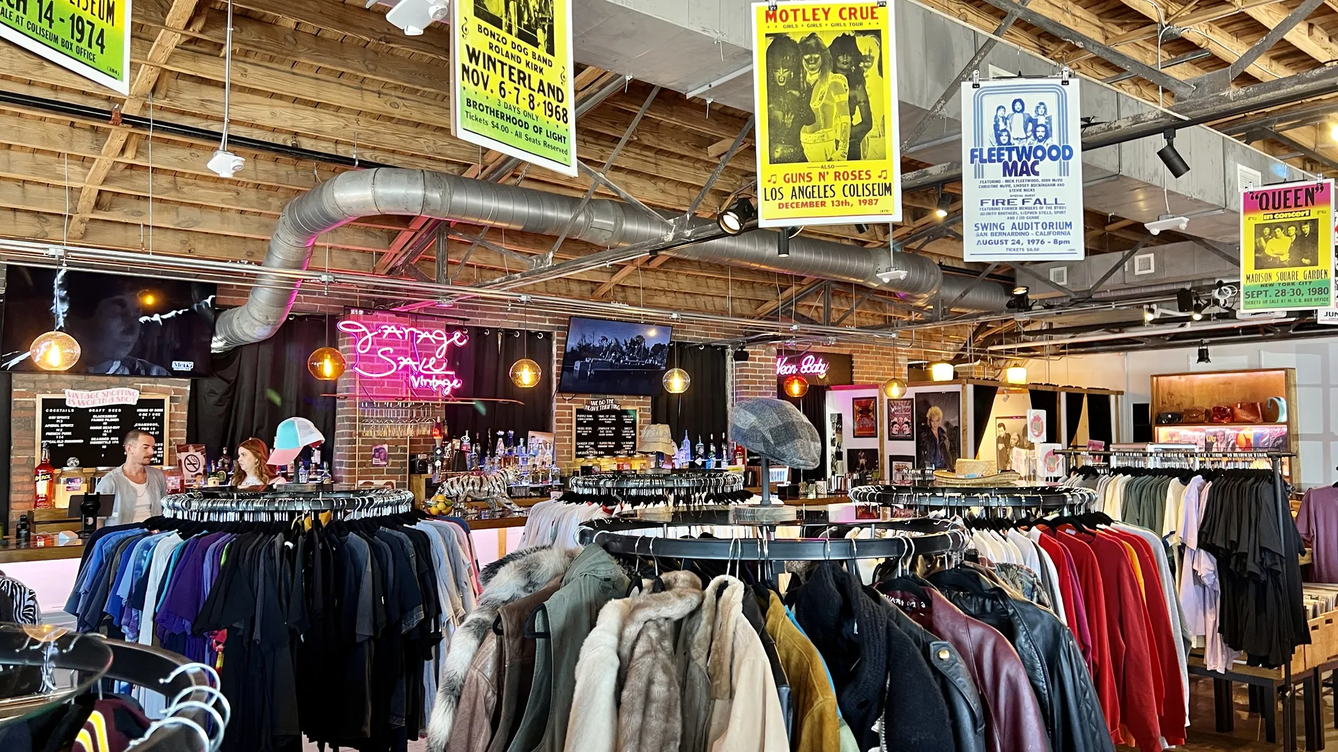 Vintage clothing store with racks of colorful jackets and shirts, neon ‘Garage Sale Vintage’ sign above a retro-style bar, exposed wooden ceiling, and hanging rock concert posters.