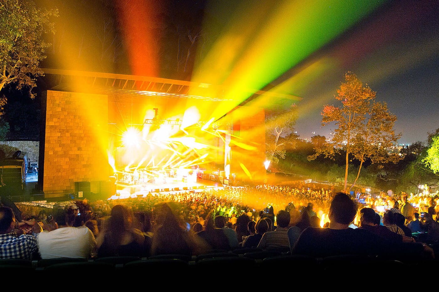 A vibrant outdoor concert at night with colorful stage lights beaming over a large crowd at an amphitheater venue.