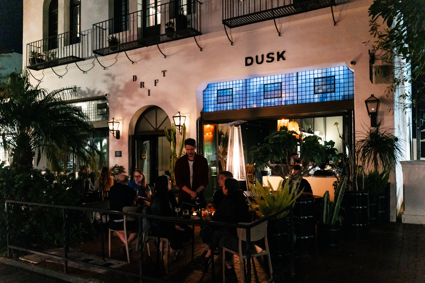 Nighttime outdoor dining at Drift and Dusk restaurant with patrons seated among lush tropical plants and warm lighting