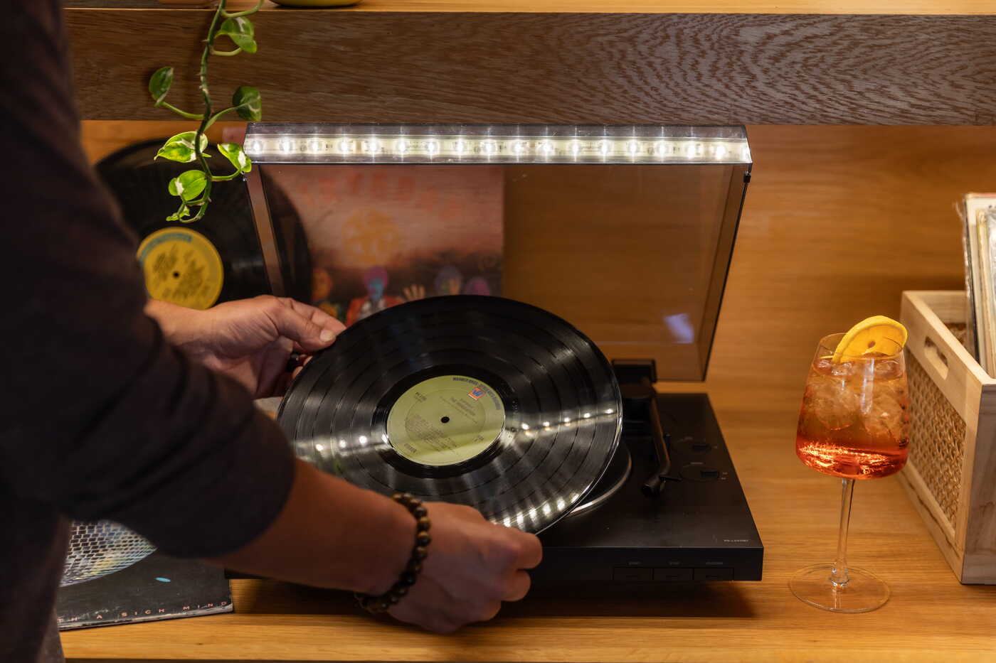 Person placing a vinyl record on a turntable beside an Aperol spritz cocktail and a crate of album collections