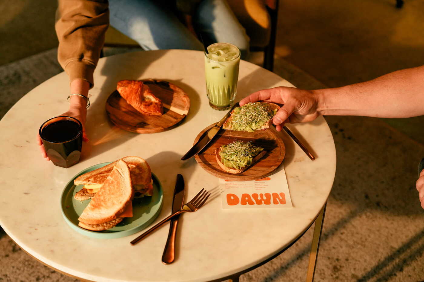 Two guests sharing avocado toast, a croissant, a sandwich, and an iced matcha latte at Dawn Coffee café