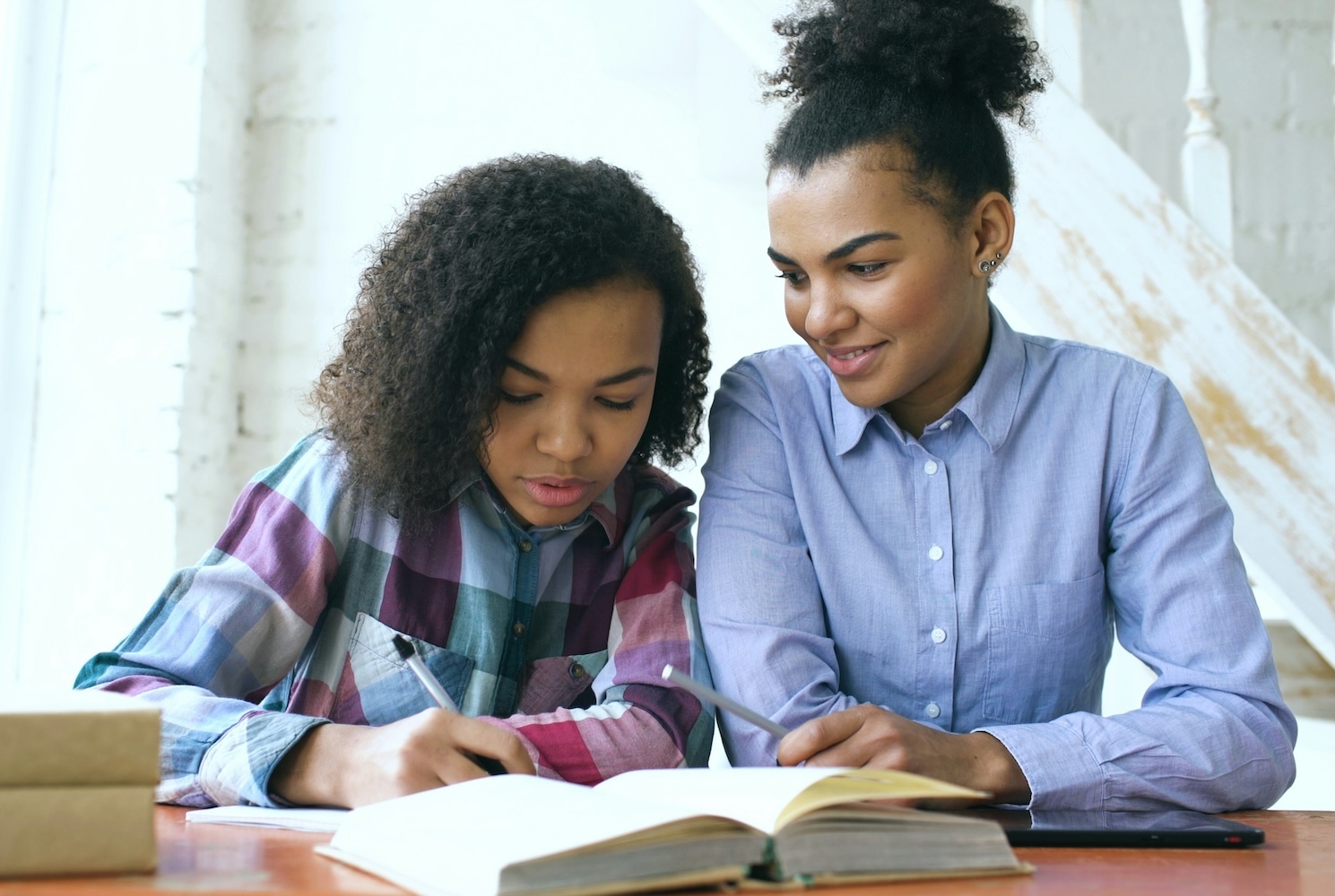 A mother looks on, smiling, as her teen daughter writes notes while looking at a hardcover book in front of them.