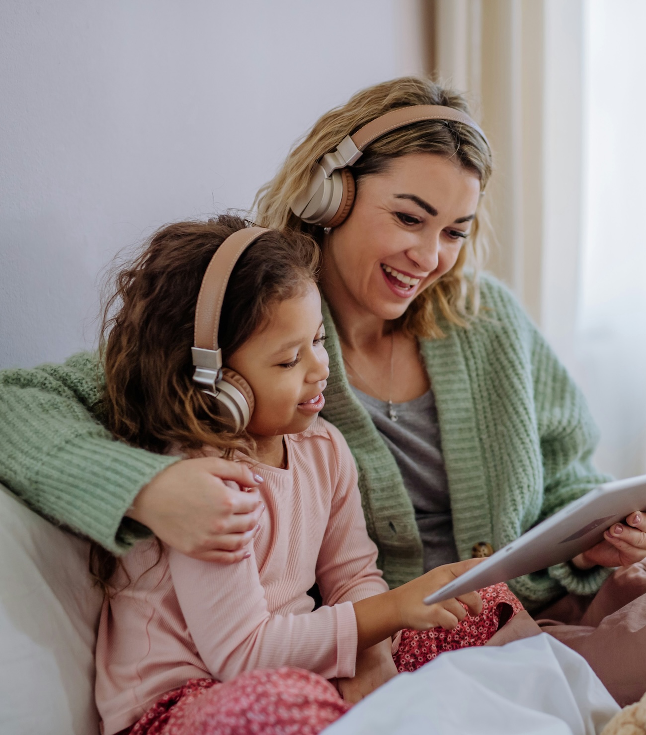 A mother and her young daughter work on a tablet together, smiling, both wearing headphones