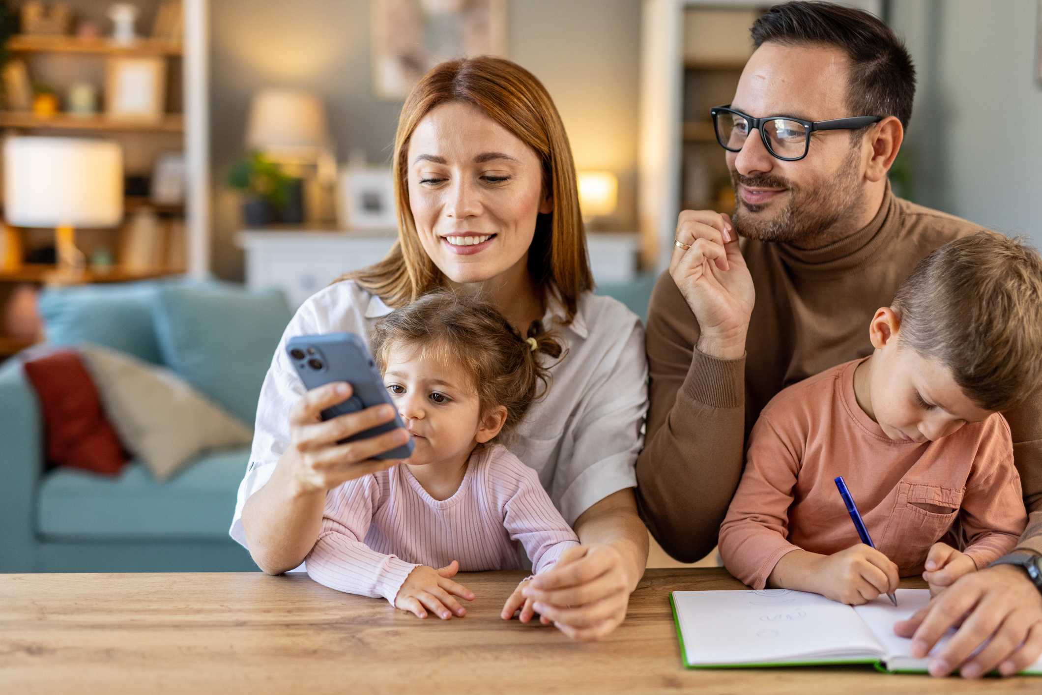 A smiling family sits at a table together, as the parents look at a phone.