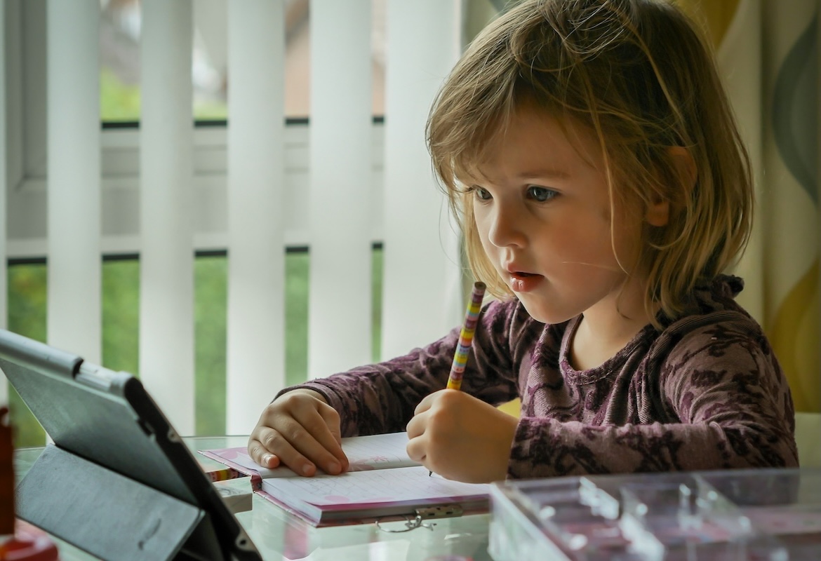 A 5 year old girl holds writes with a pencil on a small notebook, while looking at a tabled propped up in front of her.