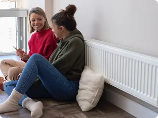 Two young women sitting on the floor by a radiator, smiling and looking at a smartphone.