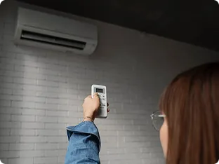 Person holding a remote control pointed at a wall-mounted air conditioner in a dimly lit room.