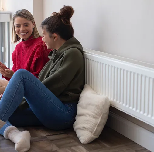 Two young women sitting on the floor by a radiator, smiling and talking with each other.