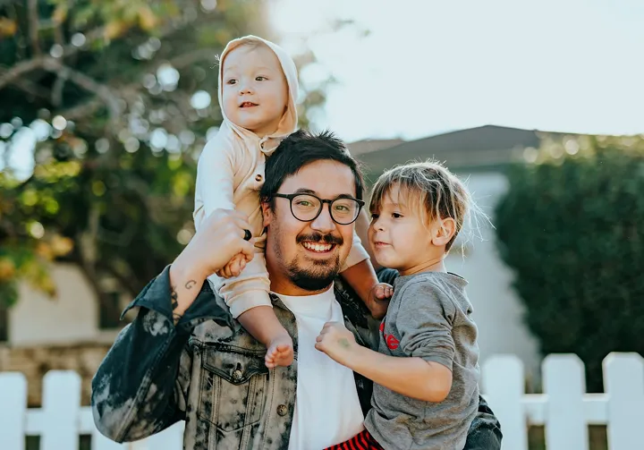 Smiling man wearing glasses holding two young children outdoors near a white fence.
