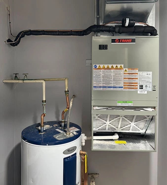 A Trane HVAC system with visible instruction labels is mounted on a wall beside a blue and white Whirlpool water heater. 