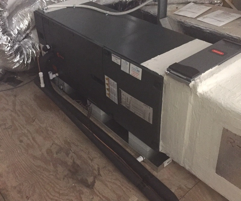 Large black air conditioning unit installed in an attic, surrounded by metallic ducts and pipes. 