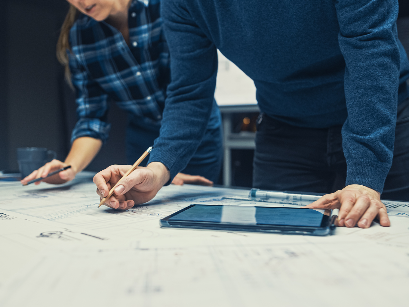 Two people leaning over a table working on architectural blueprints with a tablet and pencils.