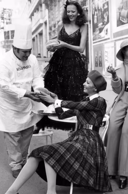 Chef handing a bread roll to a smiling woman in a plaid dress and beret seated outdoors, with a woman in a dark dress standing behind them.