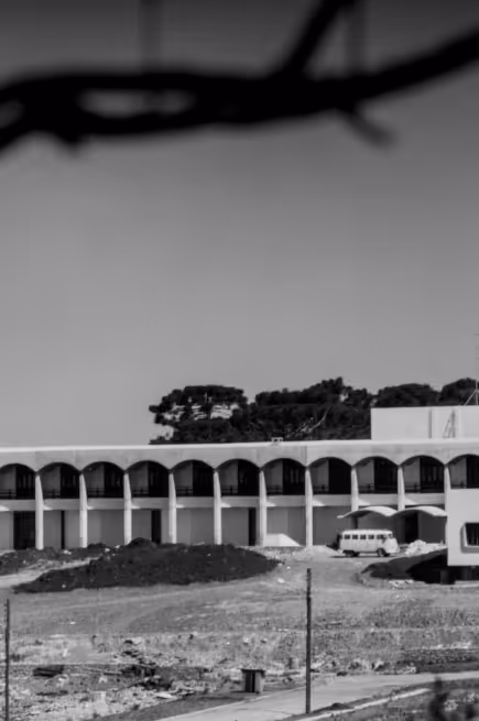 Prédio com fachada branca e arcos na parte superior, ao fundo árvores e um céu claro, com terreno aberto e um veículo estacionado na frente.