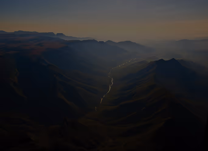 Vista aérea de uma cordilheira ao entardecer com um rio serpenteando pelo vale e neblina suave no horizonte.