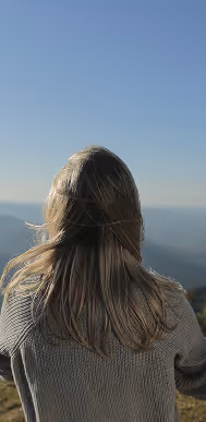 Pessoa de costas com cabelo comprido olhando para uma paisagem montanhosa sob um céu azul claro.