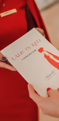 Cartão branco com texto vermelho 'LADY IN RED A WARM WELCOME' segurado por duas mãos, com pessoa vestindo roupa vermelha ao fundo.