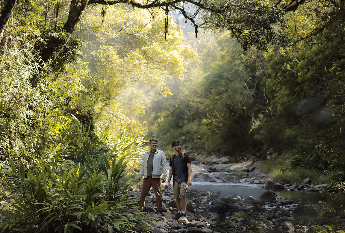 Dois homens caminhando juntos em um rio rochoso cercado por densa vegetação verde e árvores claras ao fundo.