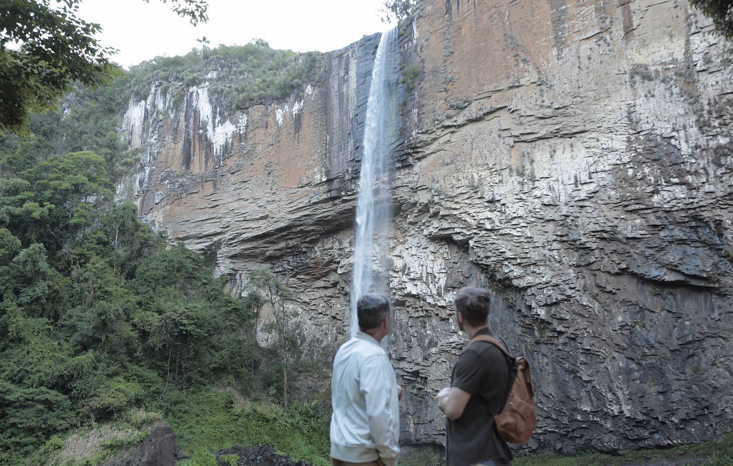 Dois homens observando uma cachoeira alta caindo de um penhasco rochoso cercado por vegetação verde.