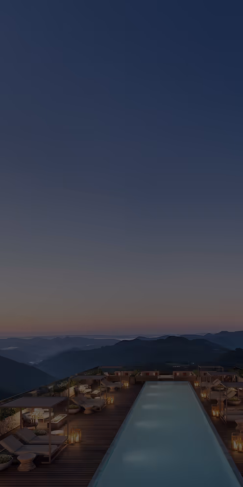 Piscina iluminada em um deck de madeira com espreguiçadeiras e lanternas ao entardecer, com montanhas ao fundo.