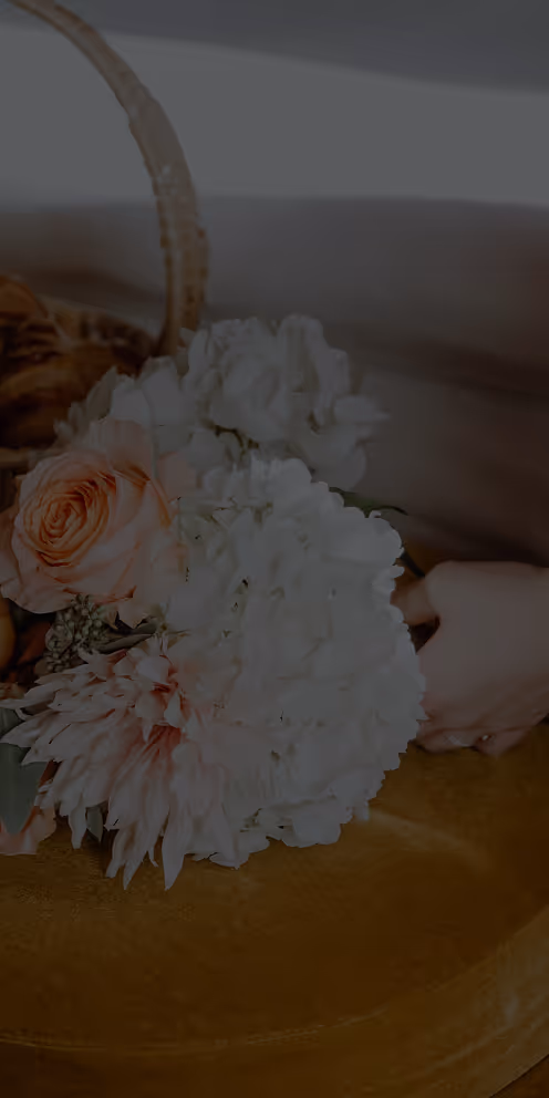 Mãos segurando um buquê de flores com rosas cor de pêssego e flores brancas em uma superfície amarela.