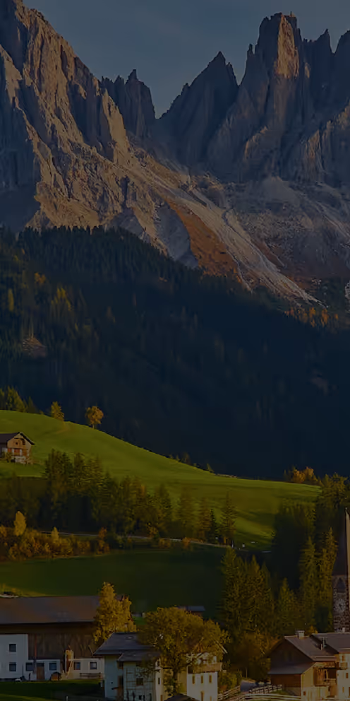 Montanhas rochosas imponentes ao fundo, com colinas verdes e casas espalhadas em uma vila tranquila.