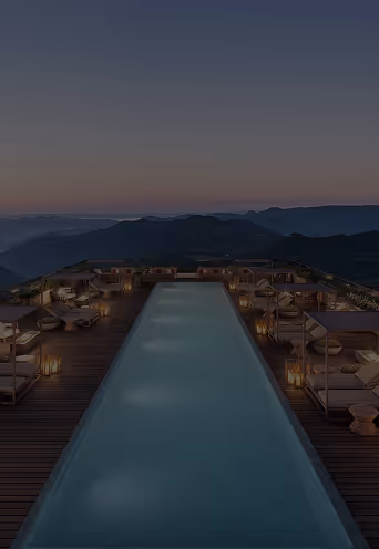 Piscina infinita iluminada à noite com espreguiçadeiras e cabanas ao redor em um terraço com vista para montanhas ao pôr do sol.